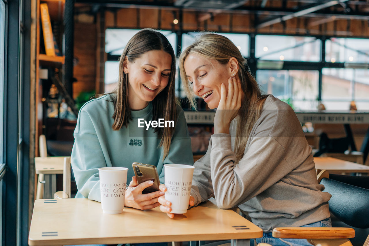 Two women sitting in a coffee house and laughing use the phone for online communication.