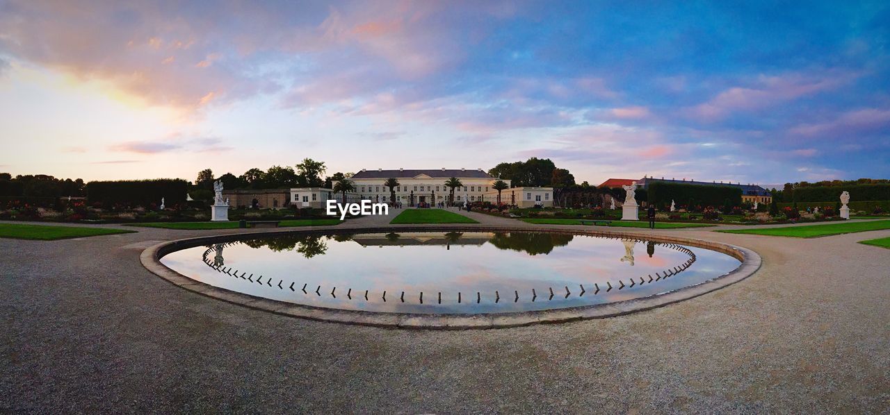 Panoramic view of palace museum and pond at herrenhausen gardens against cloudy sky