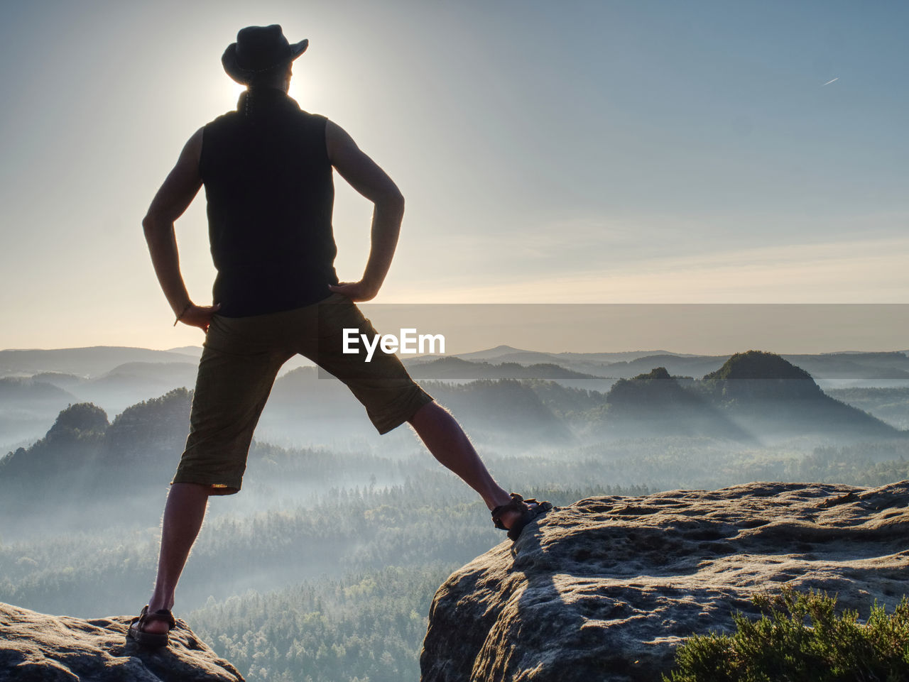 Cowboy style hiker man on cliff watching over misty morning valley to sun. man body with akimbo arms