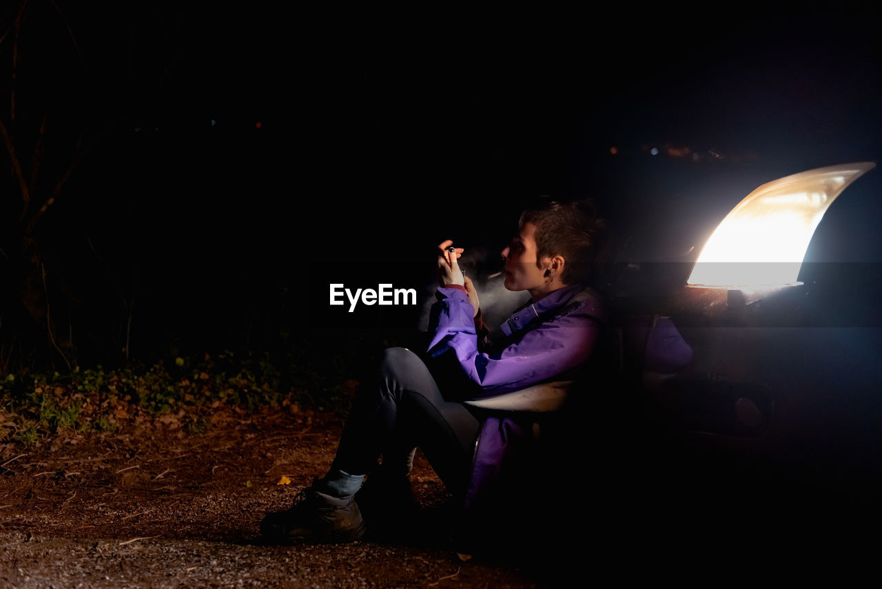 Provocative confident female in violet and vibrant orange jacket lighting cigarette while sitting alone leaning on automobile with bright headlights at dark night in spain