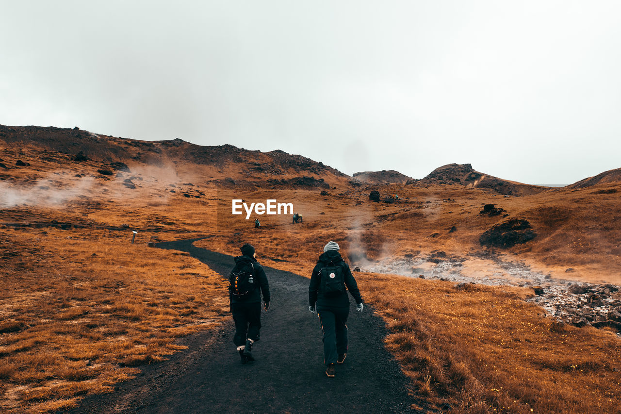 REAR VIEW OF MAN AND WOMAN WALKING ON MOUNTAIN AGAINST SKY