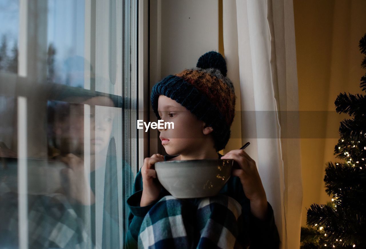 Young boy sat on a window sill with a cereal bowl looking outside