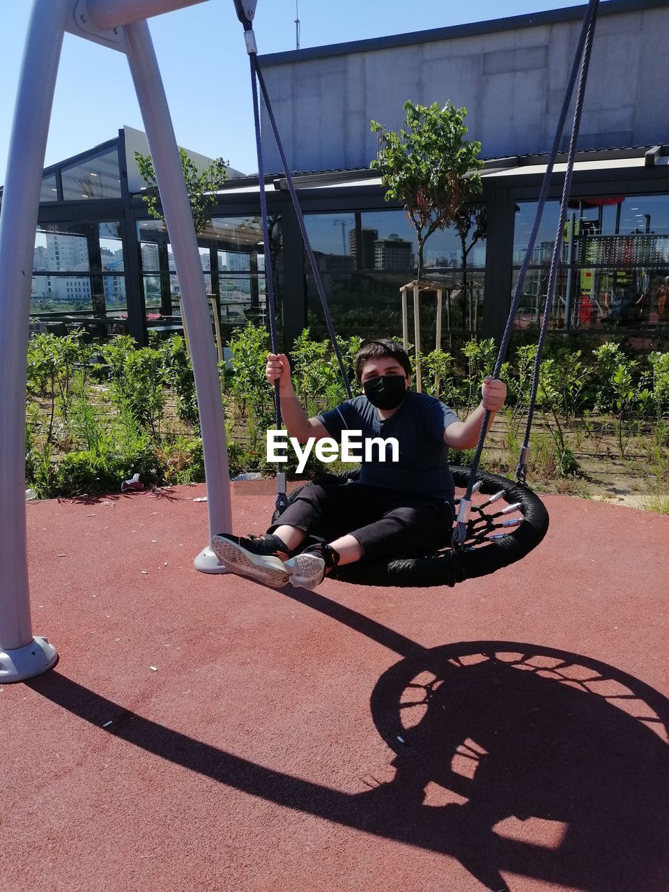 playground, full length, one person, men, lifestyles, nature, outdoor play equipment, adult, sunlight, day, shadow, city, architecture, leisure activity, sitting, built structure, plant, outdoors, sports, tree, sunny, casual clothing, sky, public space, technology