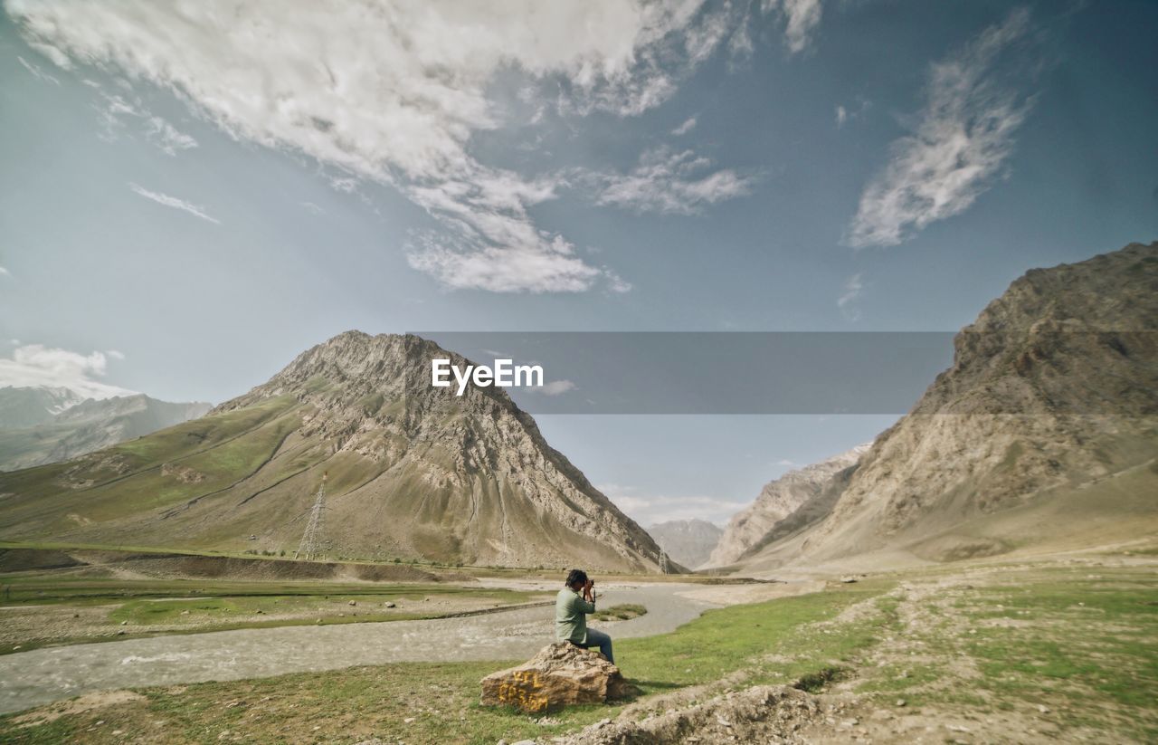 Side view of man photographing mountain while sitting on rock against sky