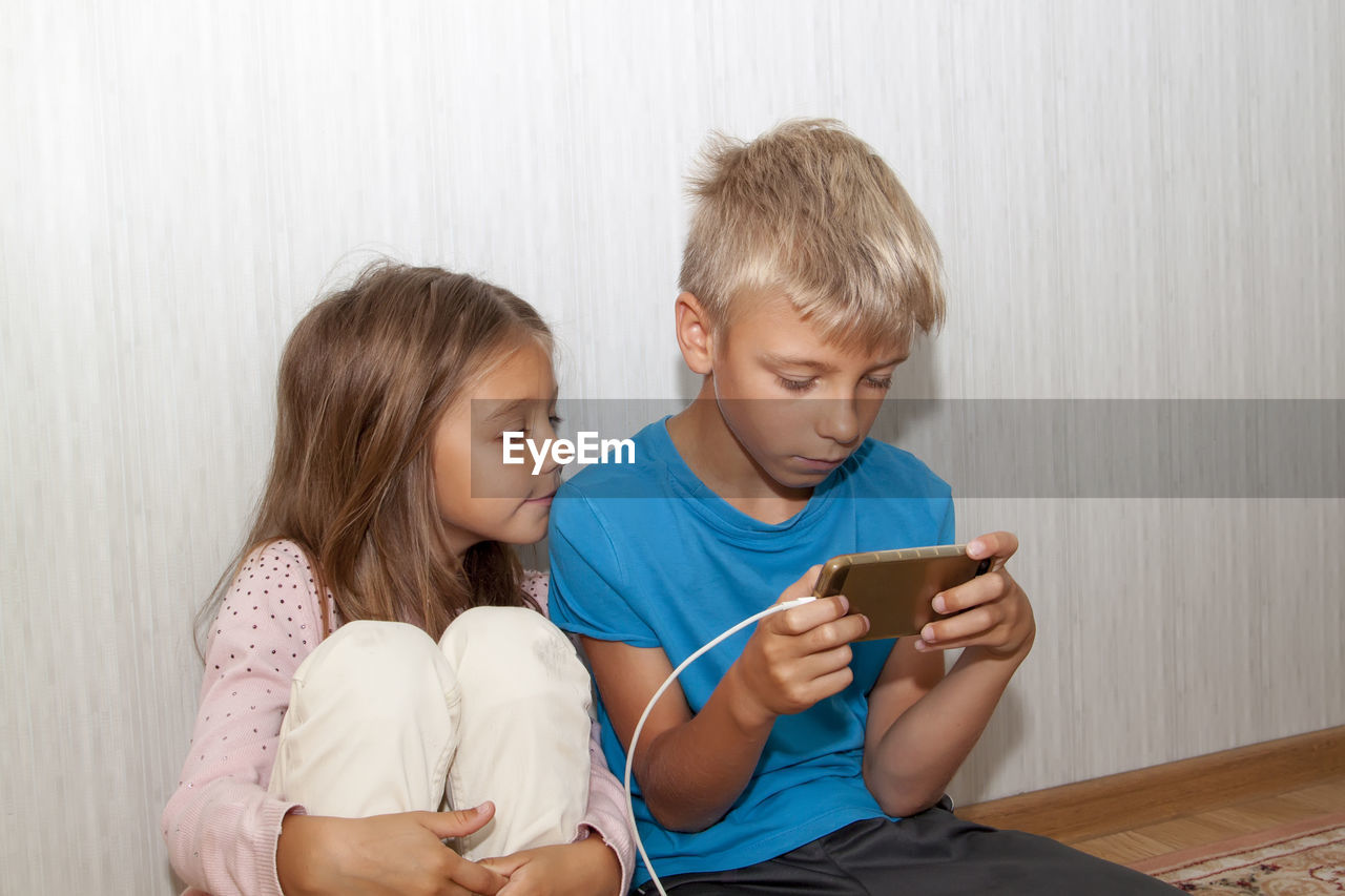 Cute boy using smart phone while sitting floor with sister at home