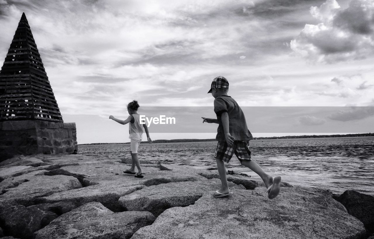Boy and girl walking at rocky beach against cloudy sky