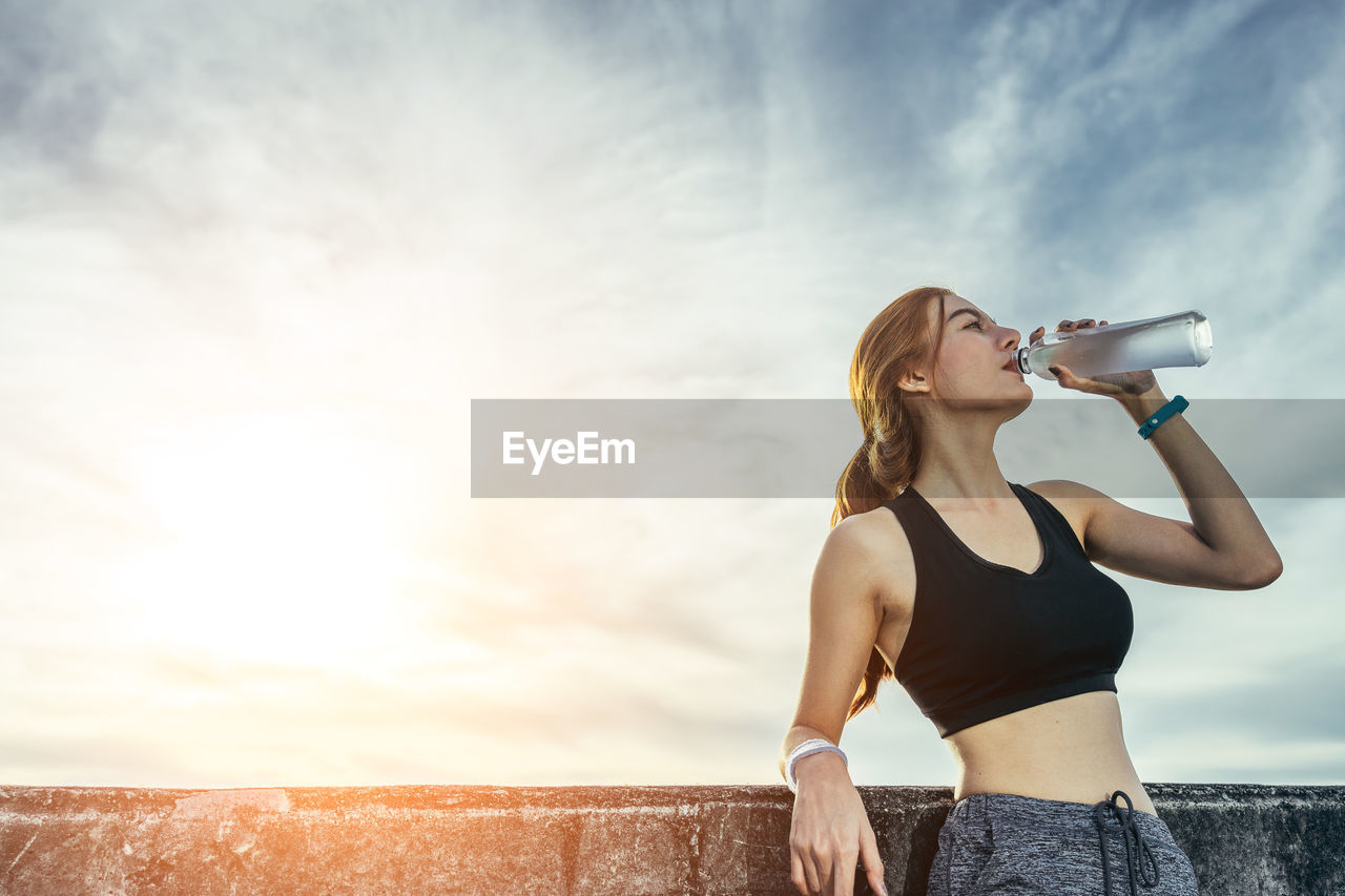 Athlete woman drinking water from bottle while standing by lake against sky