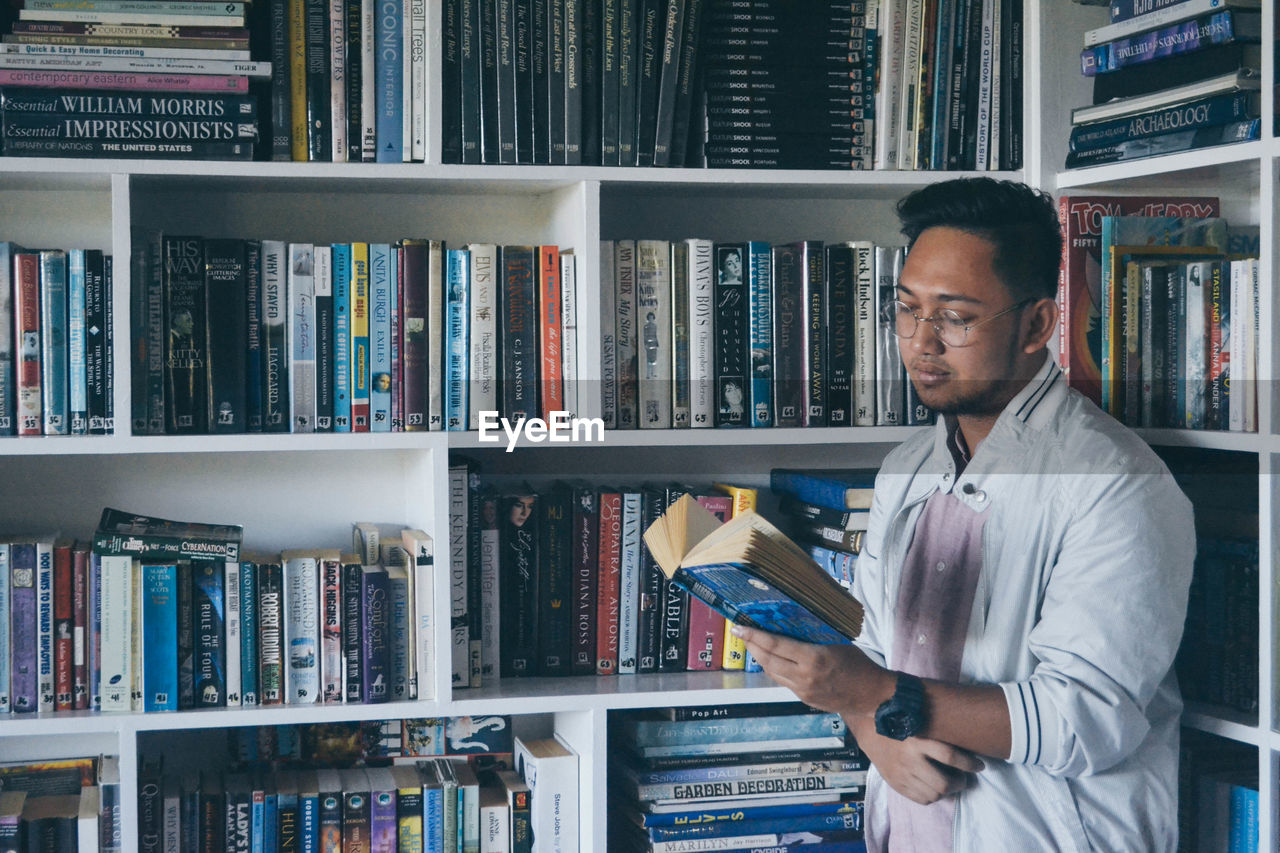 Young man reading book against shelves