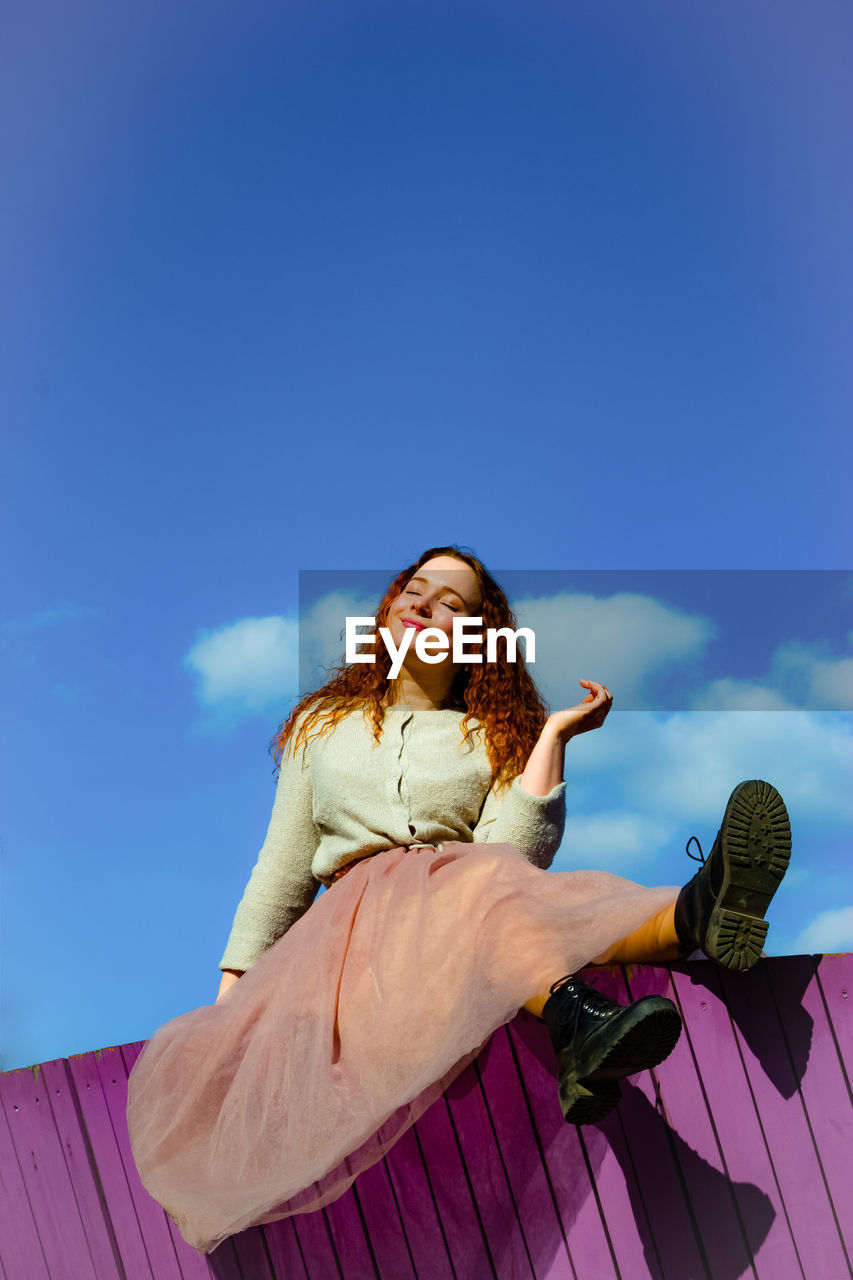 Low angle view of smiling young woman sitting on retaining wall against sky