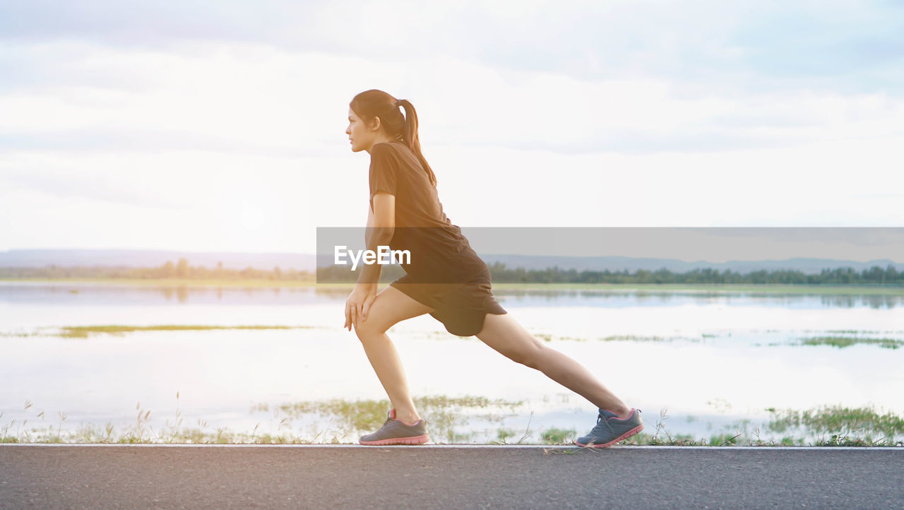 Young woman stretching on road against lake and cloudy sky