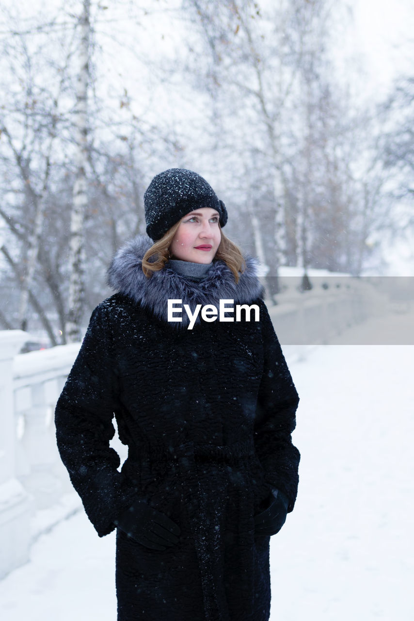 Young beautiful woman in black faux fur coat with gray fluffy hoodis walking in winter snowy park.