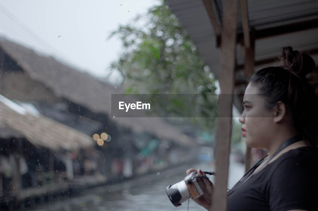 Side view of young woman holding camera during rainfall