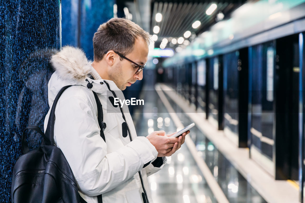 Handsome adult man in white jacket with glasses waiting subway train. killing time in mobile phone