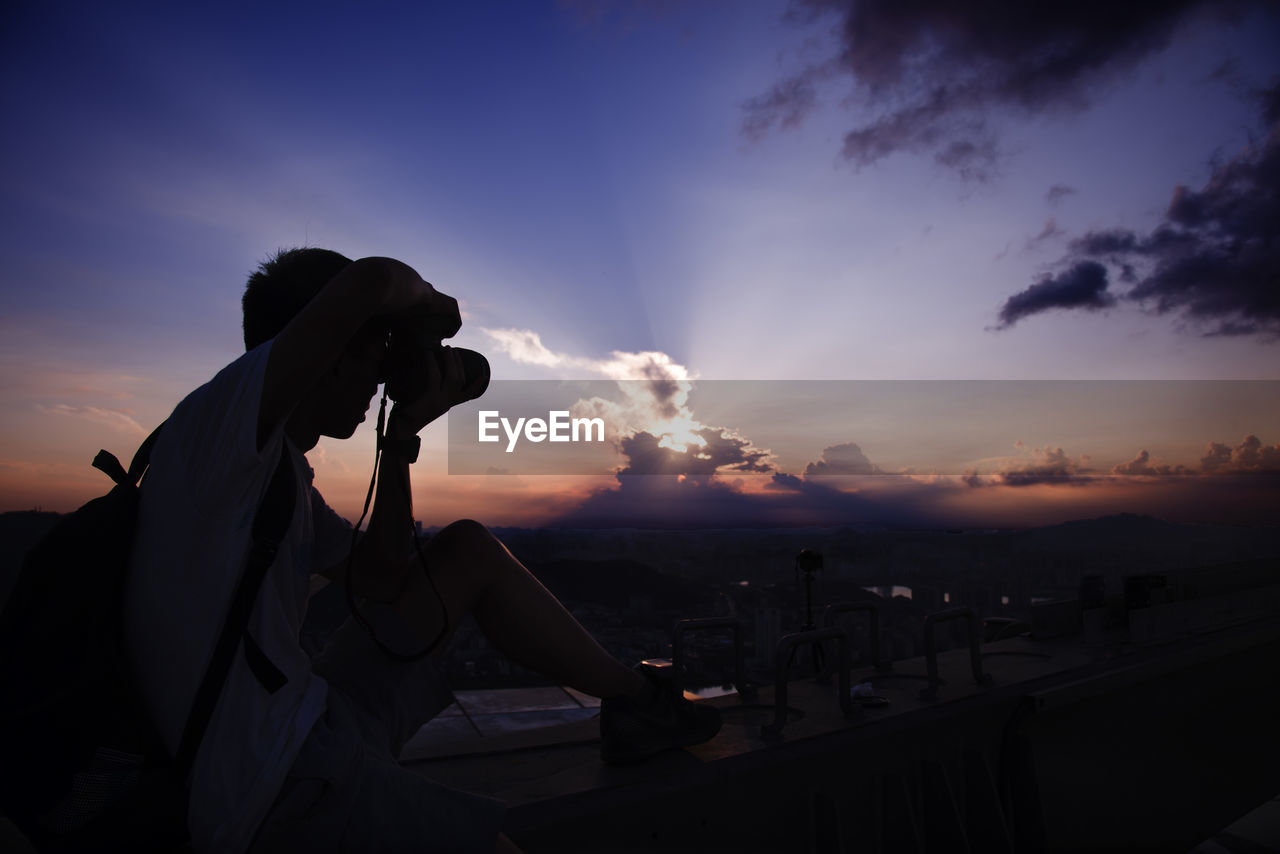 SILHOUETTE MAN PHOTOGRAPHING AGAINST SKY AT SUNSET