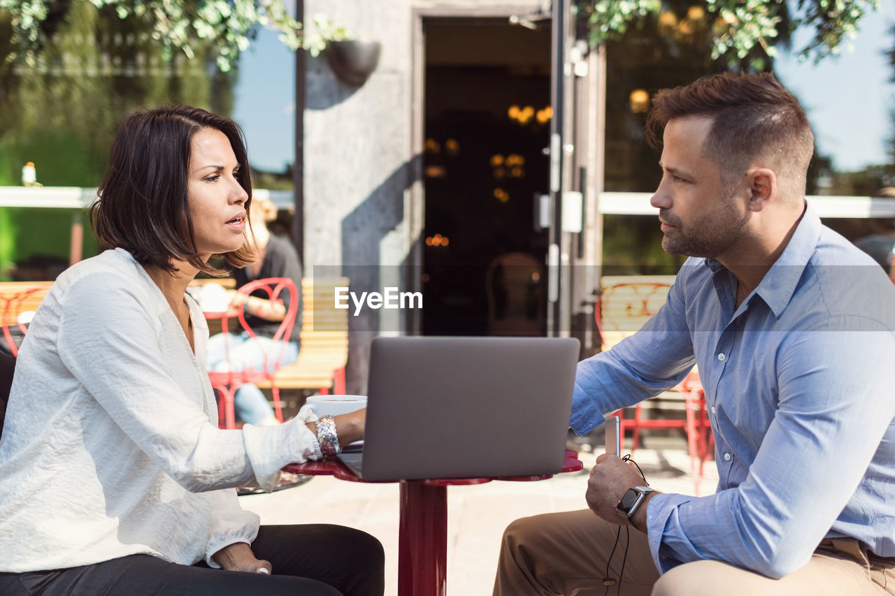 Side view of business colleagues discussing over laptop at sidewalk cafe