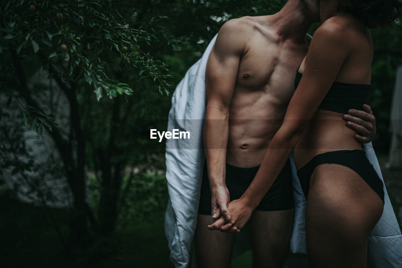 Midsection of shirtless man and woman standing against trees