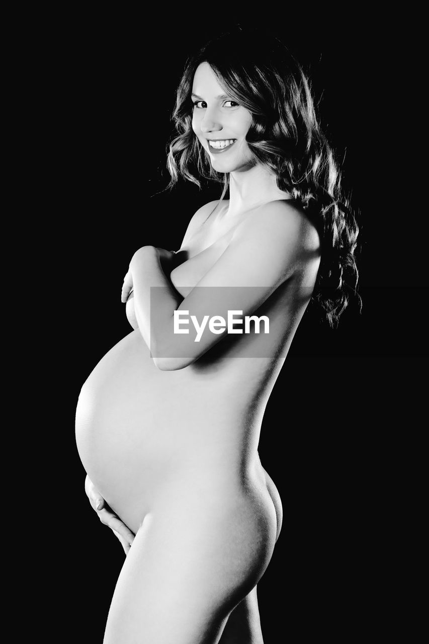 Portrait of pregnant naked woman standing against black background