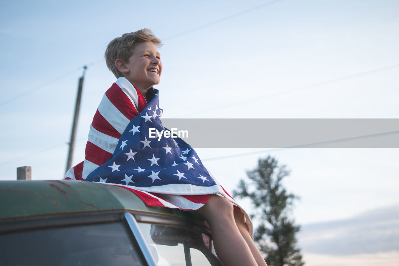 Happy boy with american flag sitting on vehicle against sky during sunset