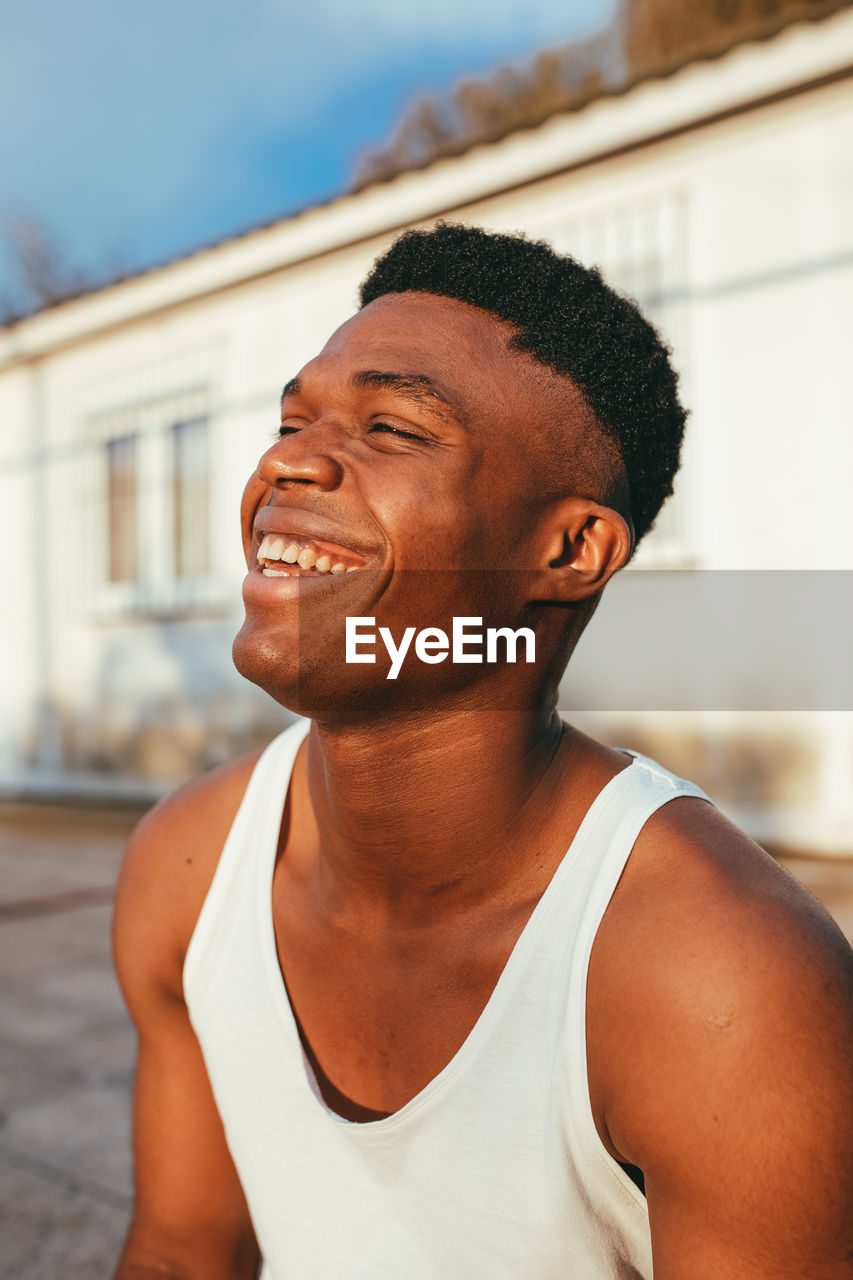 Happy african american male in undershirt with modern haircut looking forward against building in sunlight