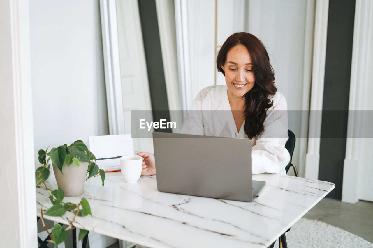 Smiling brunette woman with long hair in white cardigan working on laptop in bright modern office