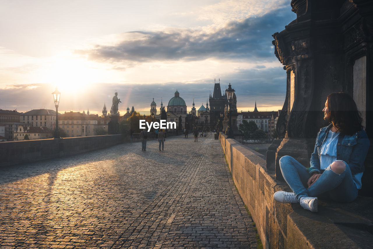 Woman sitting at charles bridge against sky during sunset