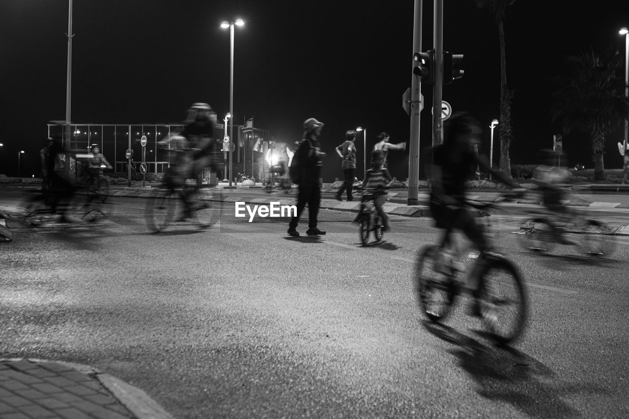 transportation, black and white, bicycle, night, street, city, motion, group of people, sports, black, monochrome photography, monochrome, road, mode of transportation, men, lifestyles, land vehicle, blurred motion, illuminated, vehicle, activity, street light, city life, adult, white, crowd, architecture, infrastructure, leisure activity, cycling, city street, riding, full length, person, large group of people, lane, women, on the move, outdoors