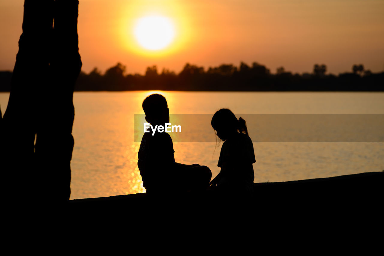 Silhouette siblings sitting at lakeshore during sunset
