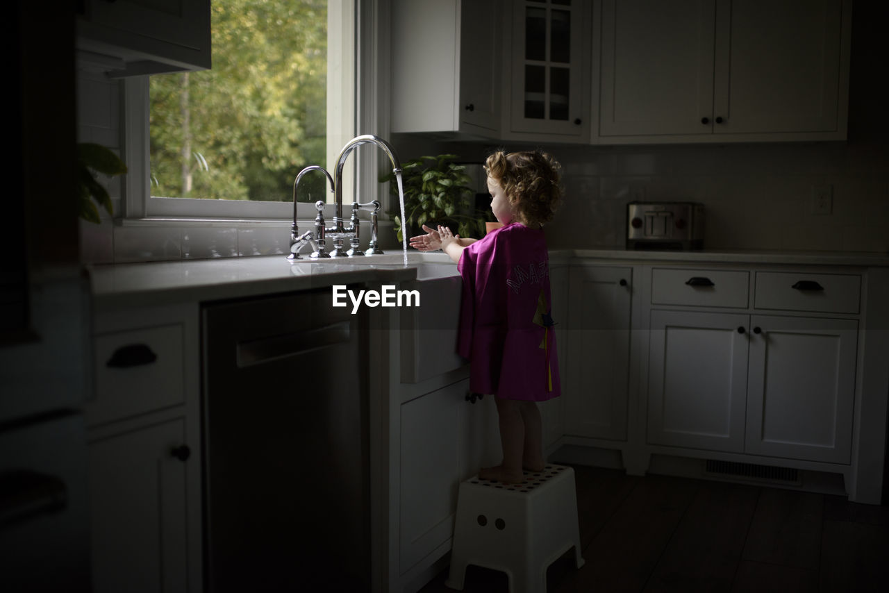 Side view of girl wearing pink cape washing hands in kitchen sink while standing on stool at home