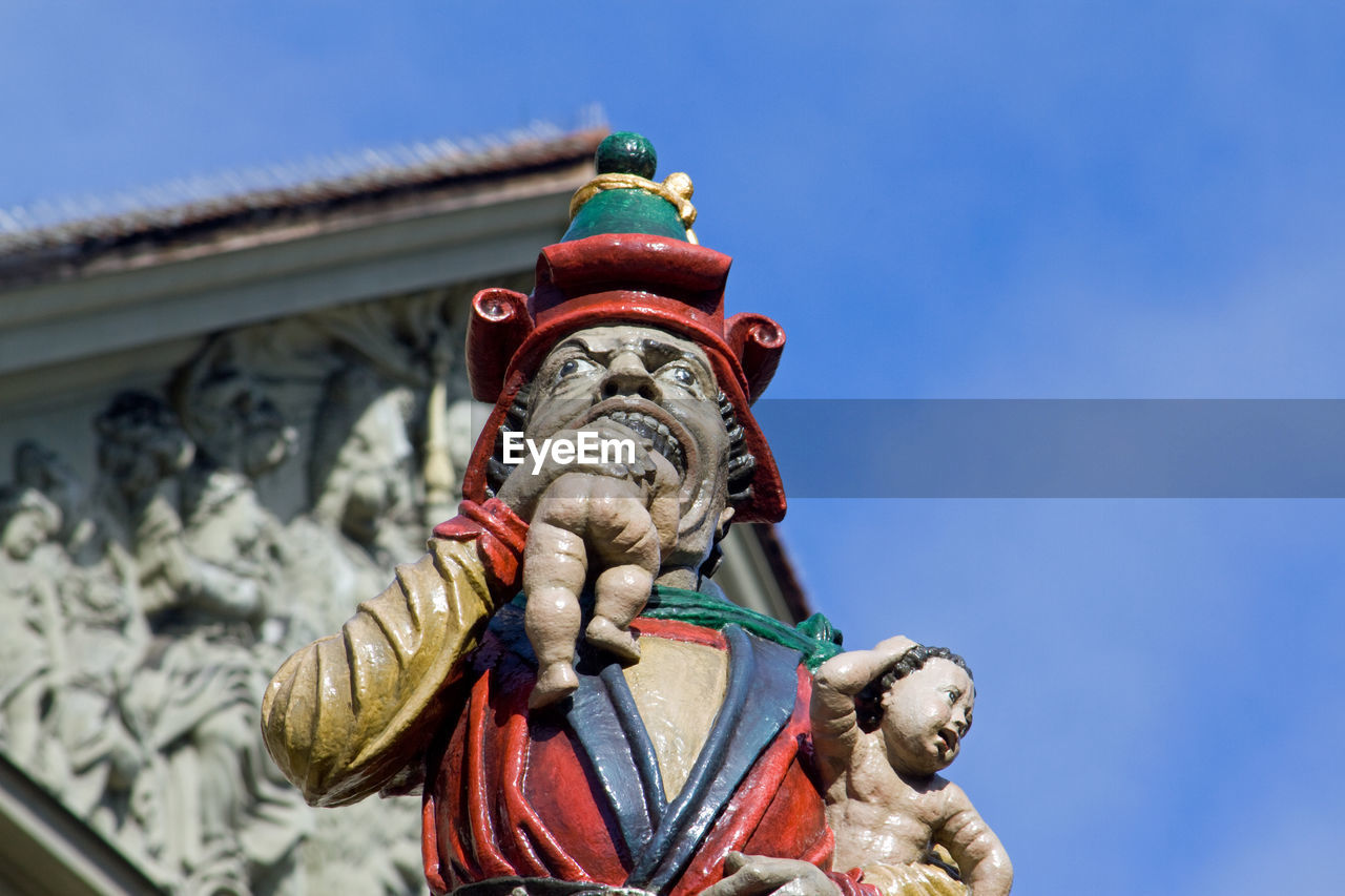 Low angle view of child-eating statue against blue sky