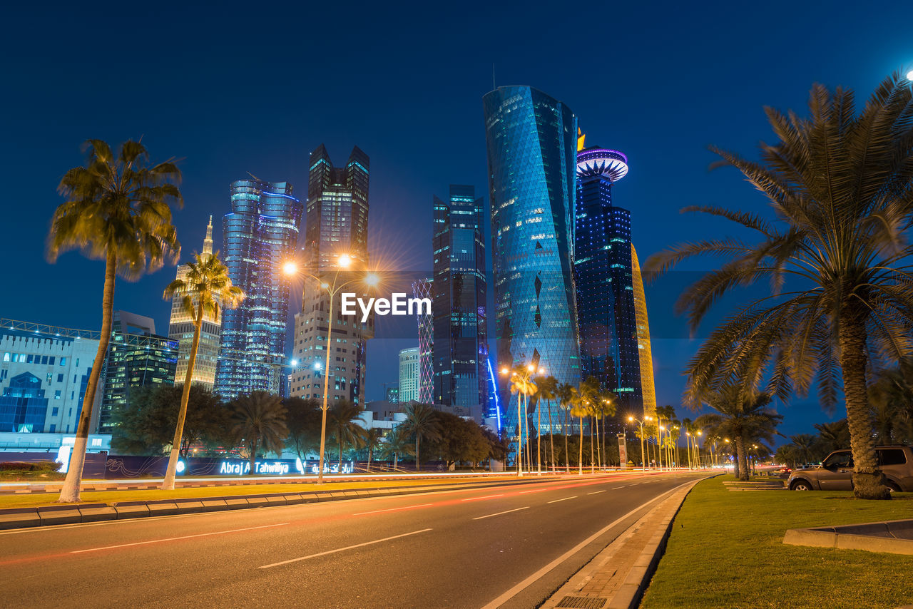 LIGHT TRAILS ON STREET AMIDST BUILDINGS AGAINST SKY AT NIGHT