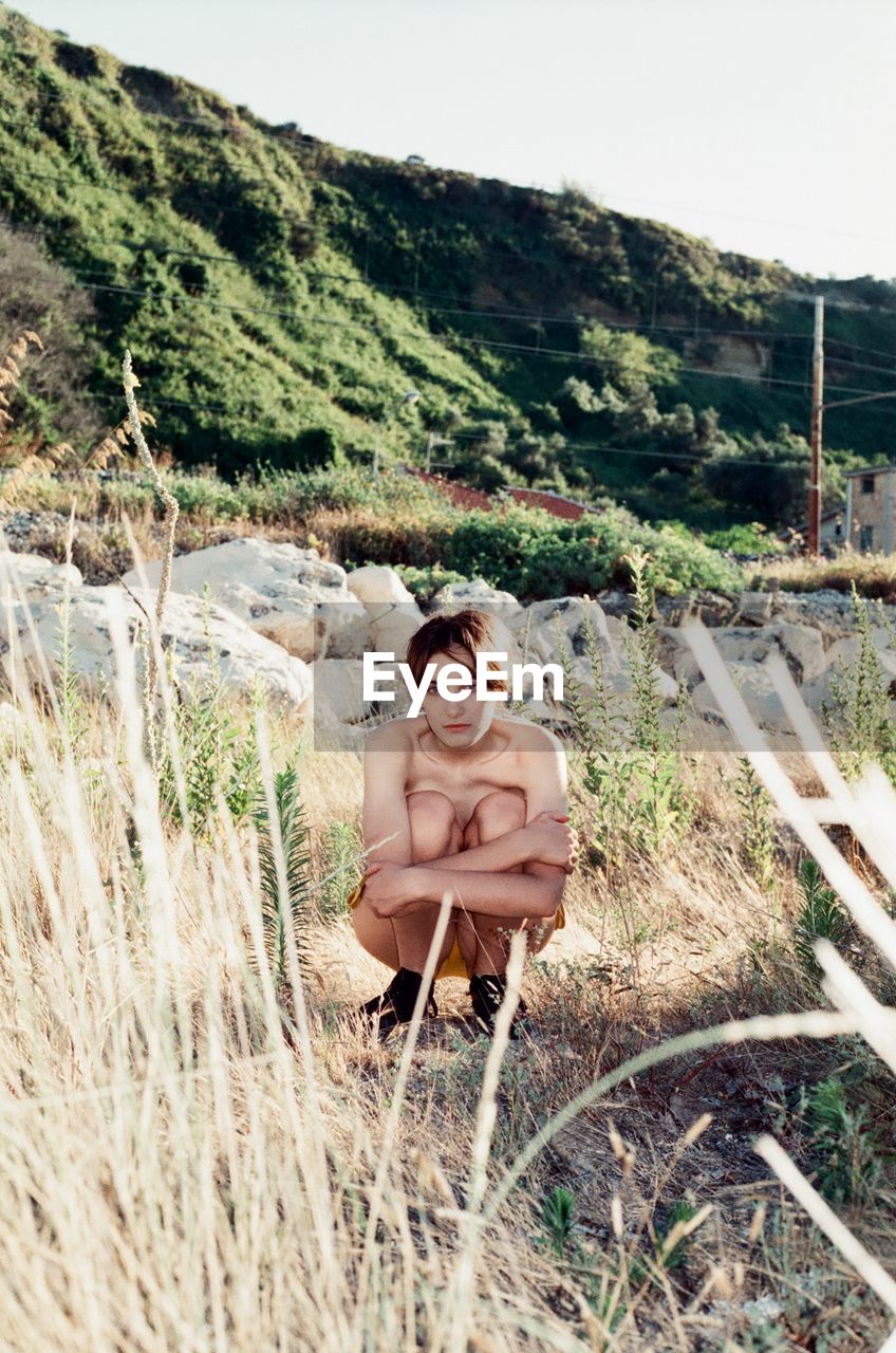 Shirtless young woman crouching on field against sky