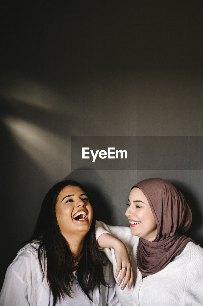 Happy young woman wearing hijab looking at friend laughing against gray background