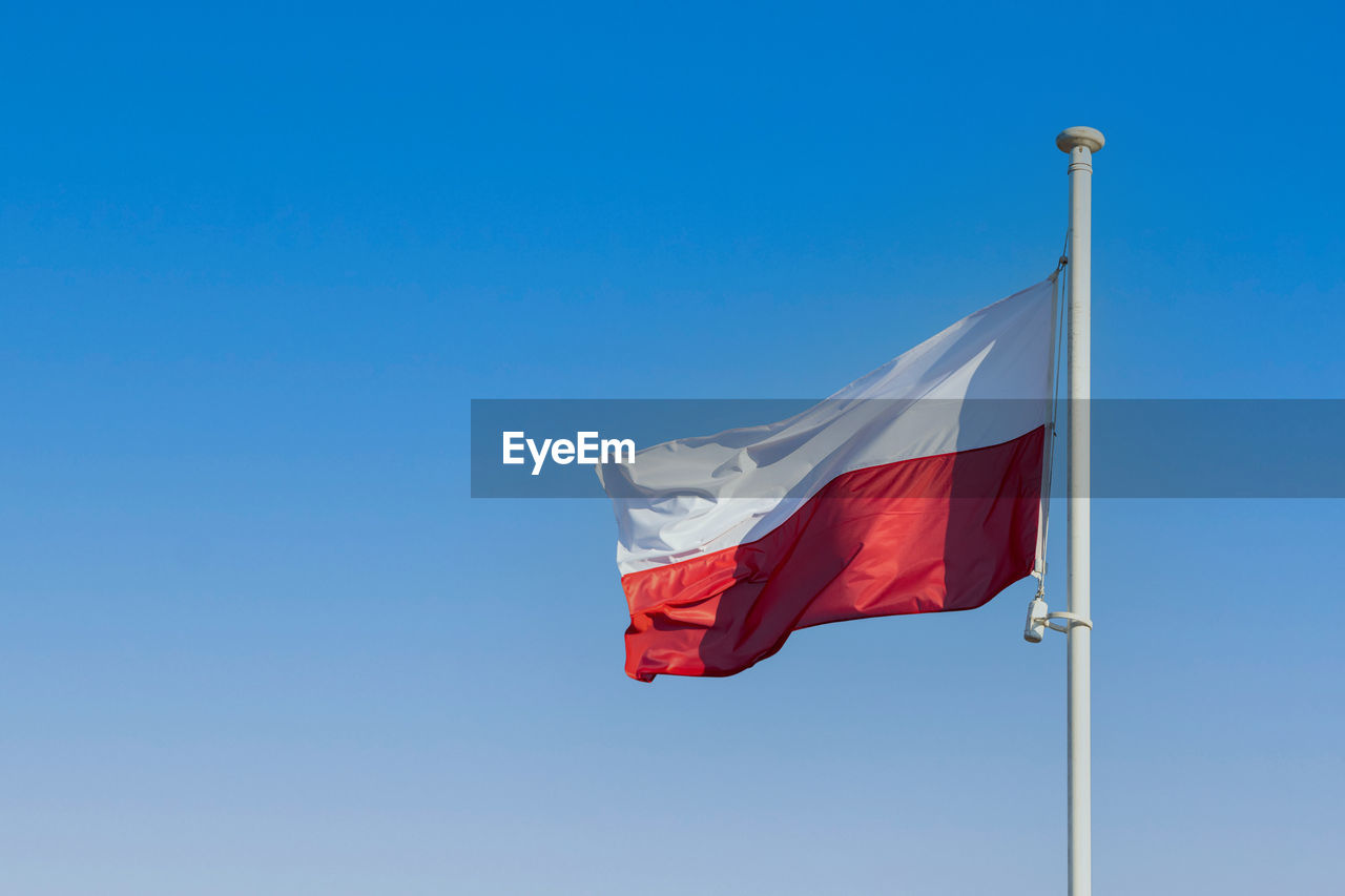 The flag of poland flying in the wind on a tall pole against a blue sky.