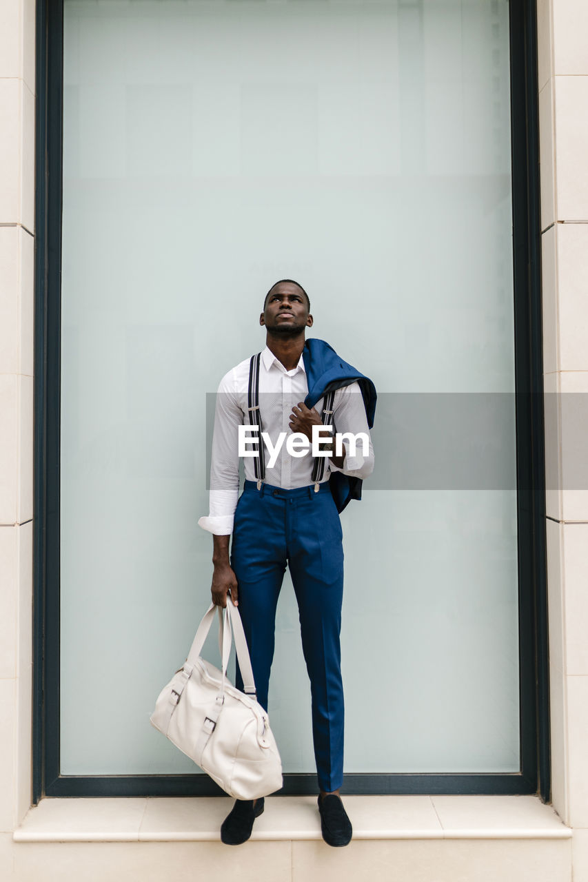 Young male professional holding suit and duffel bag while looking up against window in city