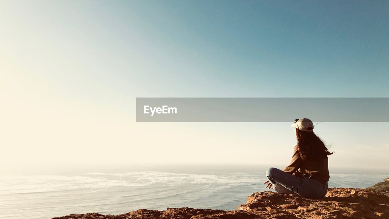 Young woman sitting on rock while looking at sea against sky