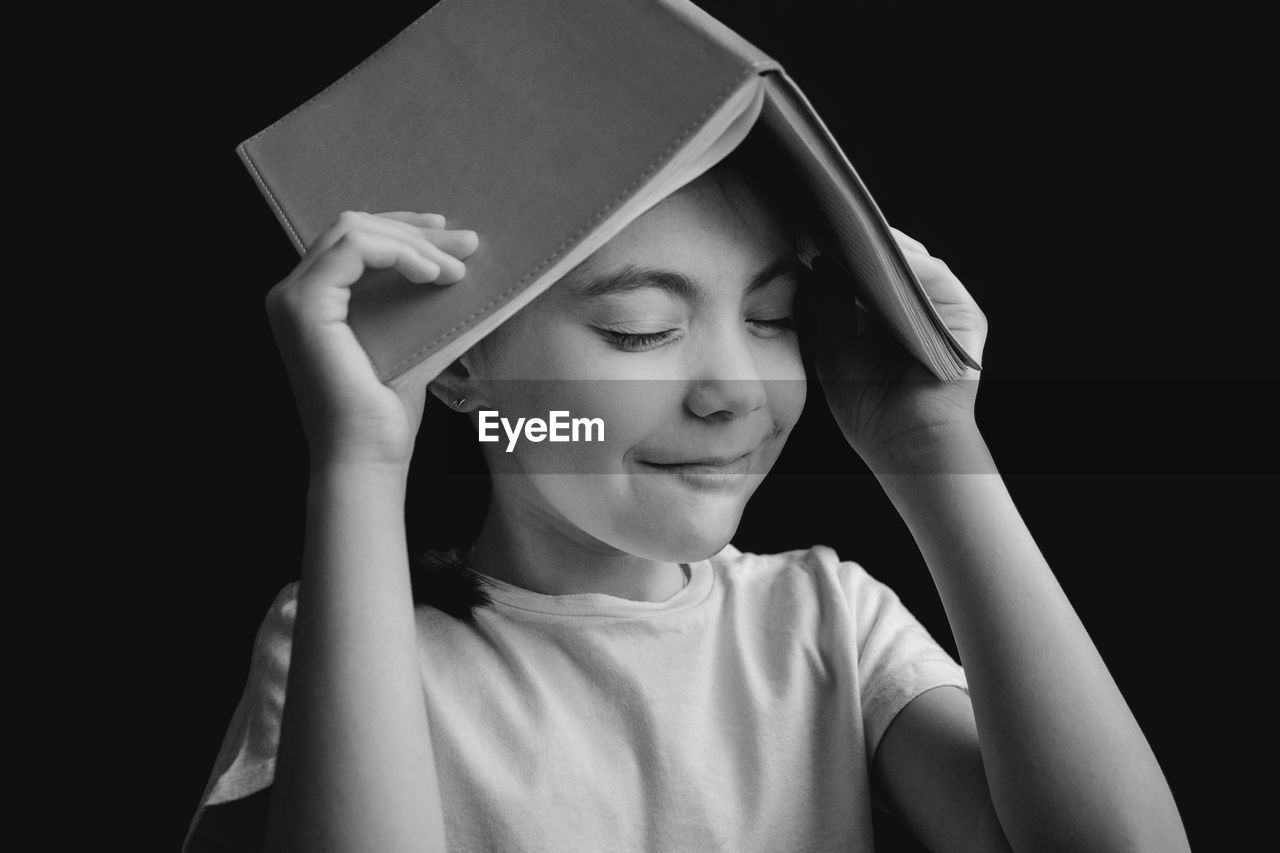 Close-up of smiling girl with eyes closed holding book on head against black background