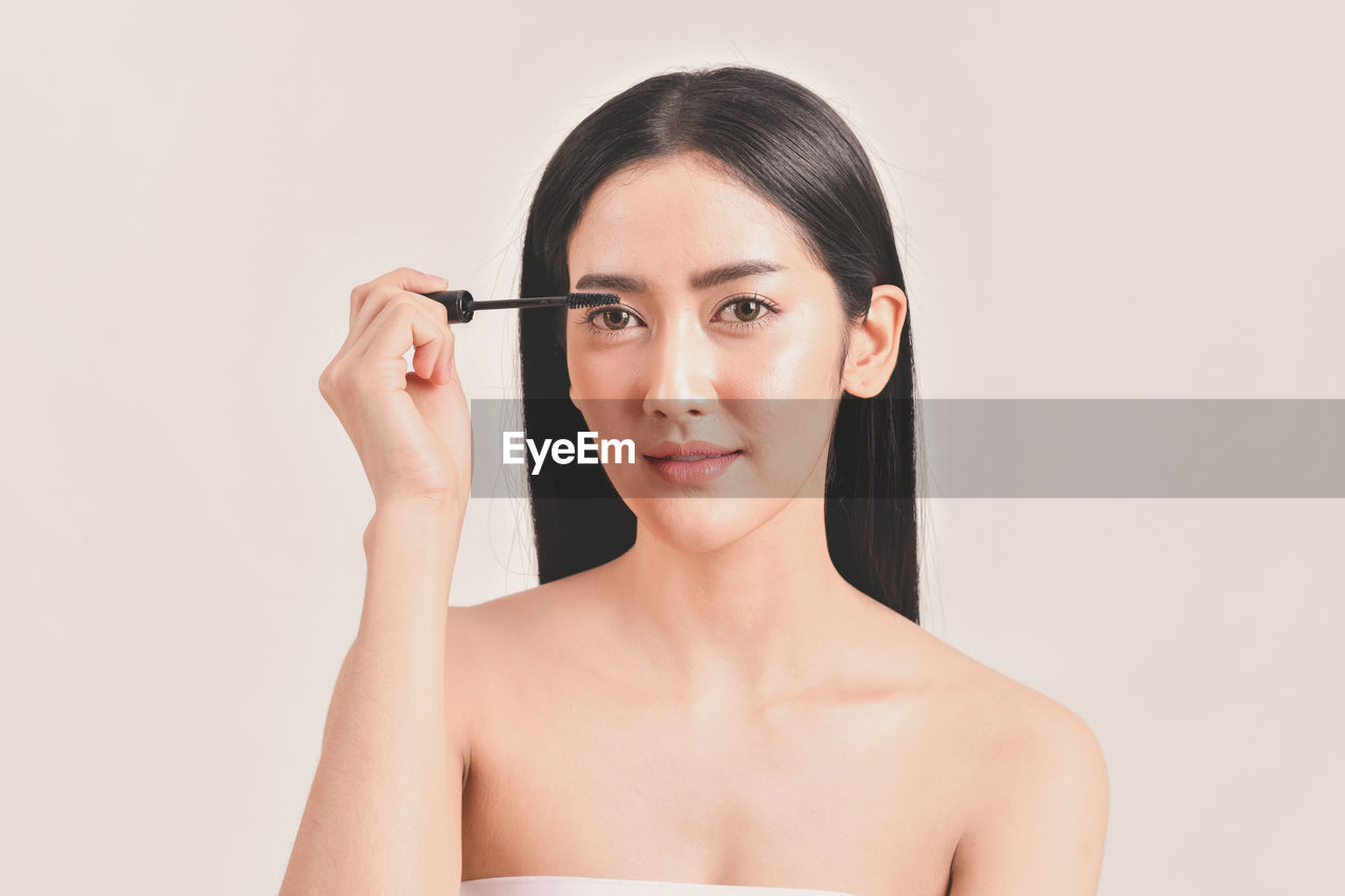 Portrait of beautiful young woman applying mascara against white background