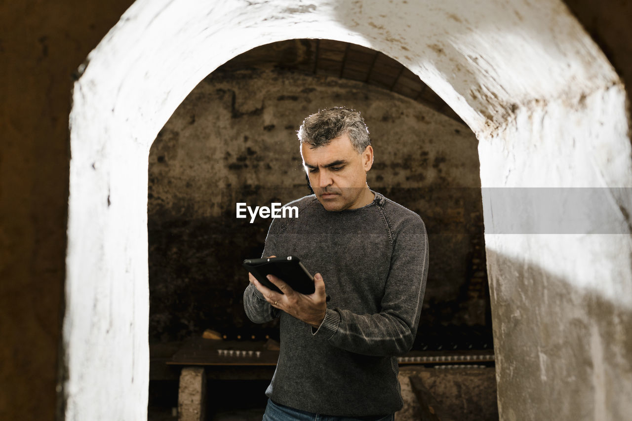 Mature man using digital tablet while standing at archway in cellar