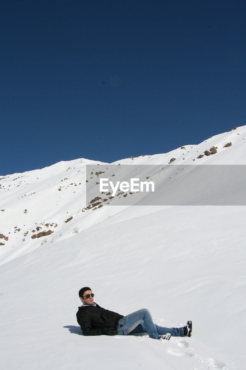 Full length of man lying down on snow covered landscape against clear blue sky