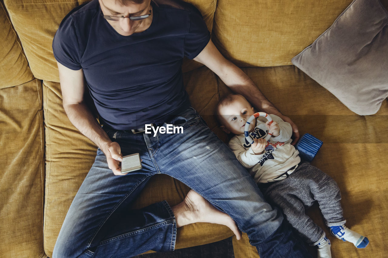 Baby boy lying on couch besides his father using smartphone