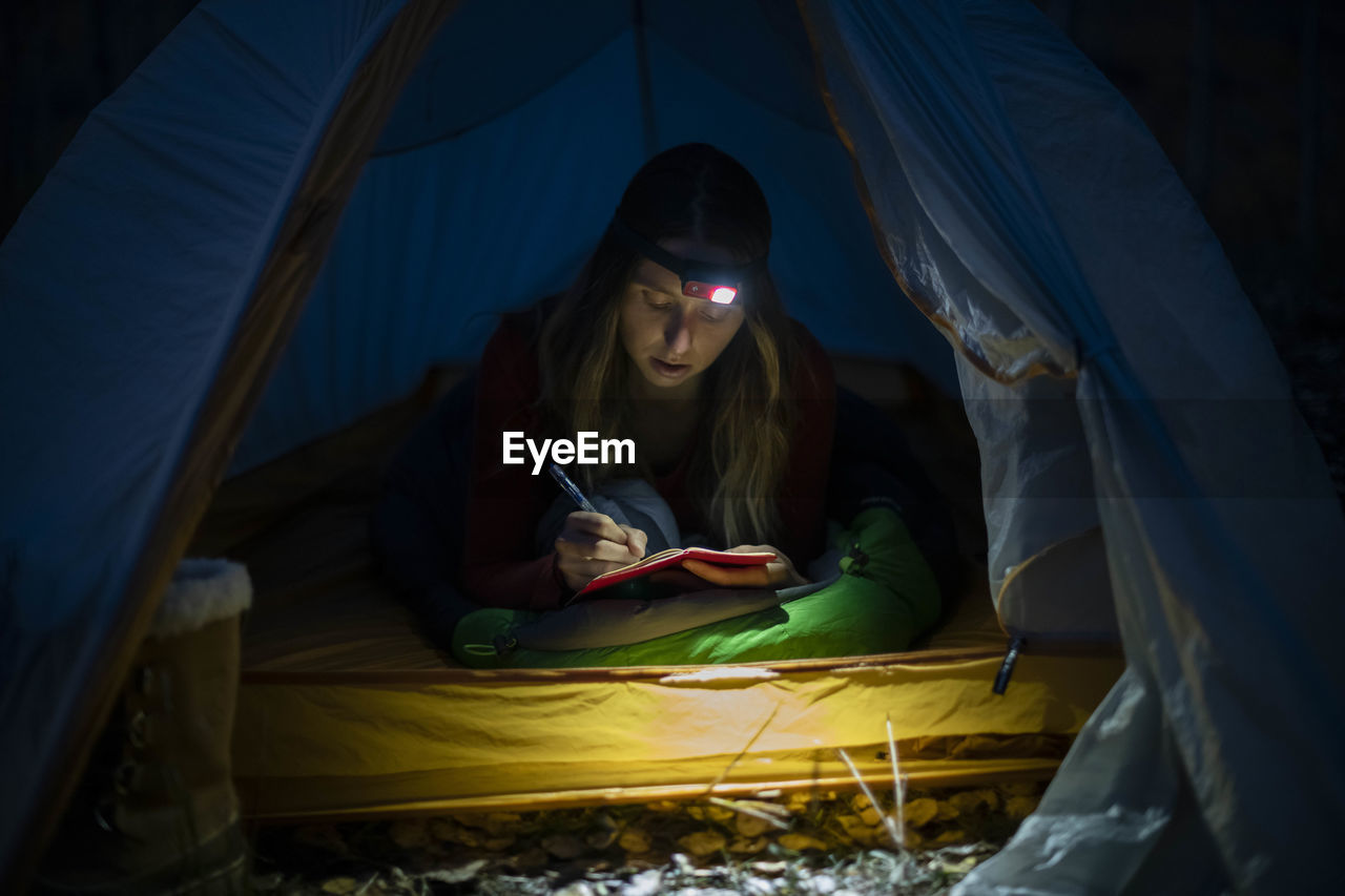Young woman writing in diary in tent at night