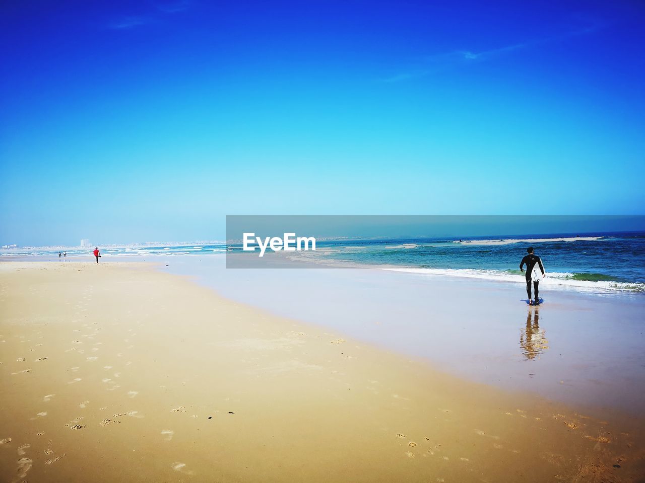 Rear view of surfer standing on shore at beach against blue sky