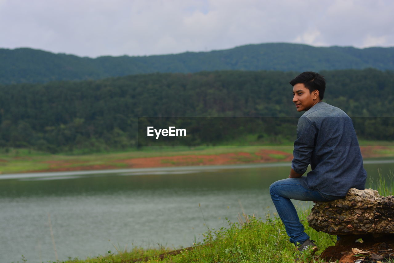 Smiling young man sitting by lake against mountain