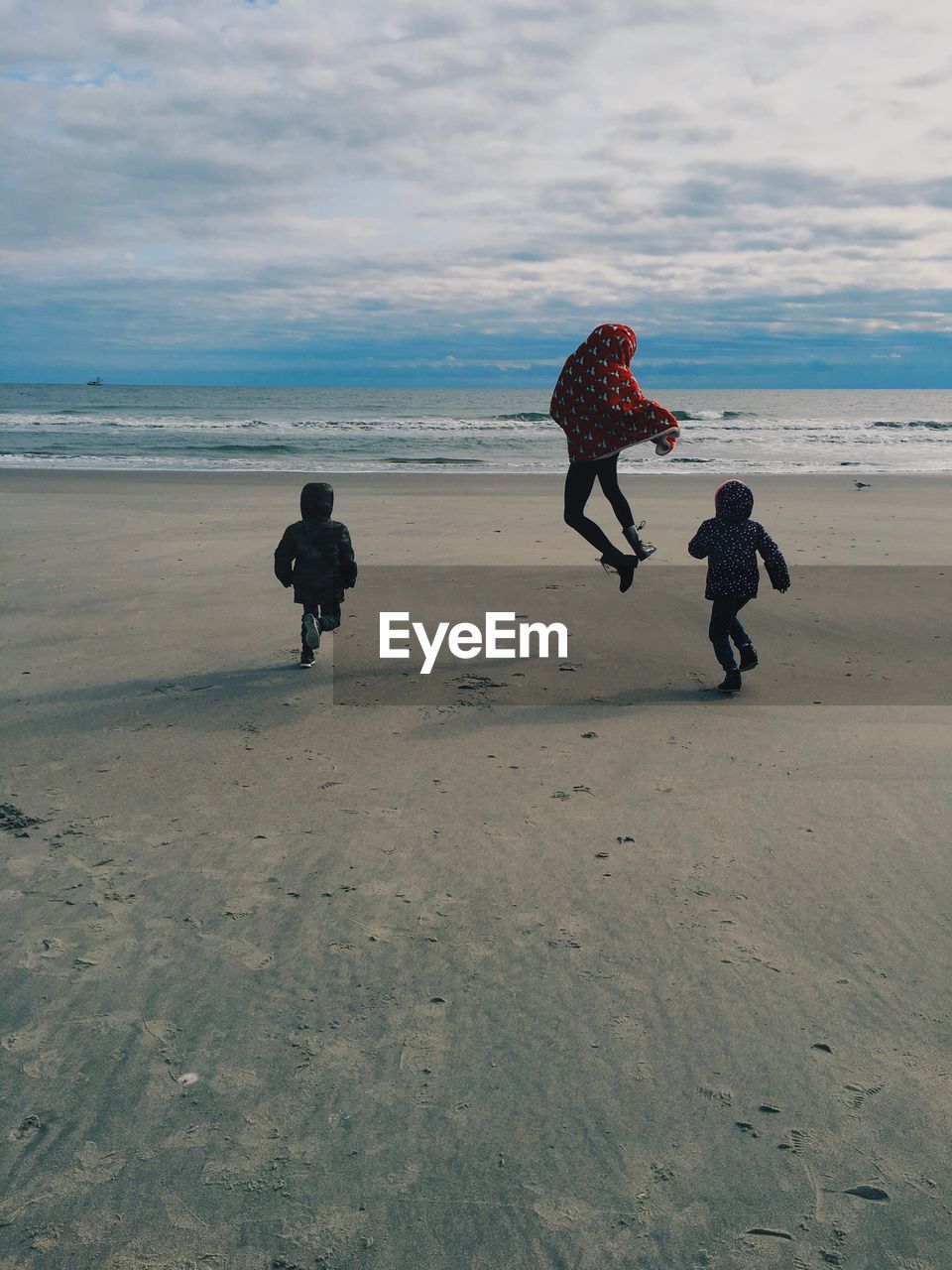 Rear view of woman jumping amidst boys at beach against sky