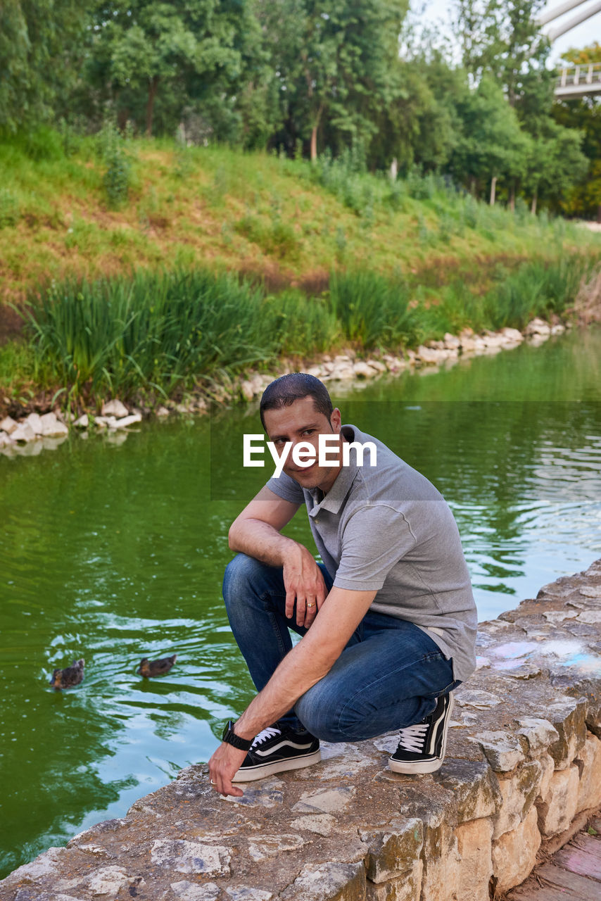 water, one person, sitting, full length, adult, men, nature, plant, lake, leisure activity, tree, casual clothing, day, rock, lifestyles, young adult, outdoors, portrait, looking at camera, smiling, relaxation, beauty in nature, tranquility, crouching, front view, emotion