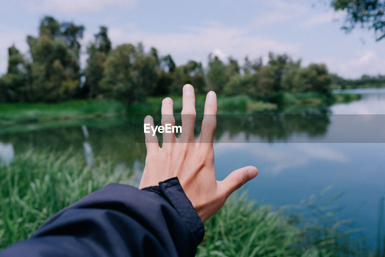 Cropped hand reaching lake against trees