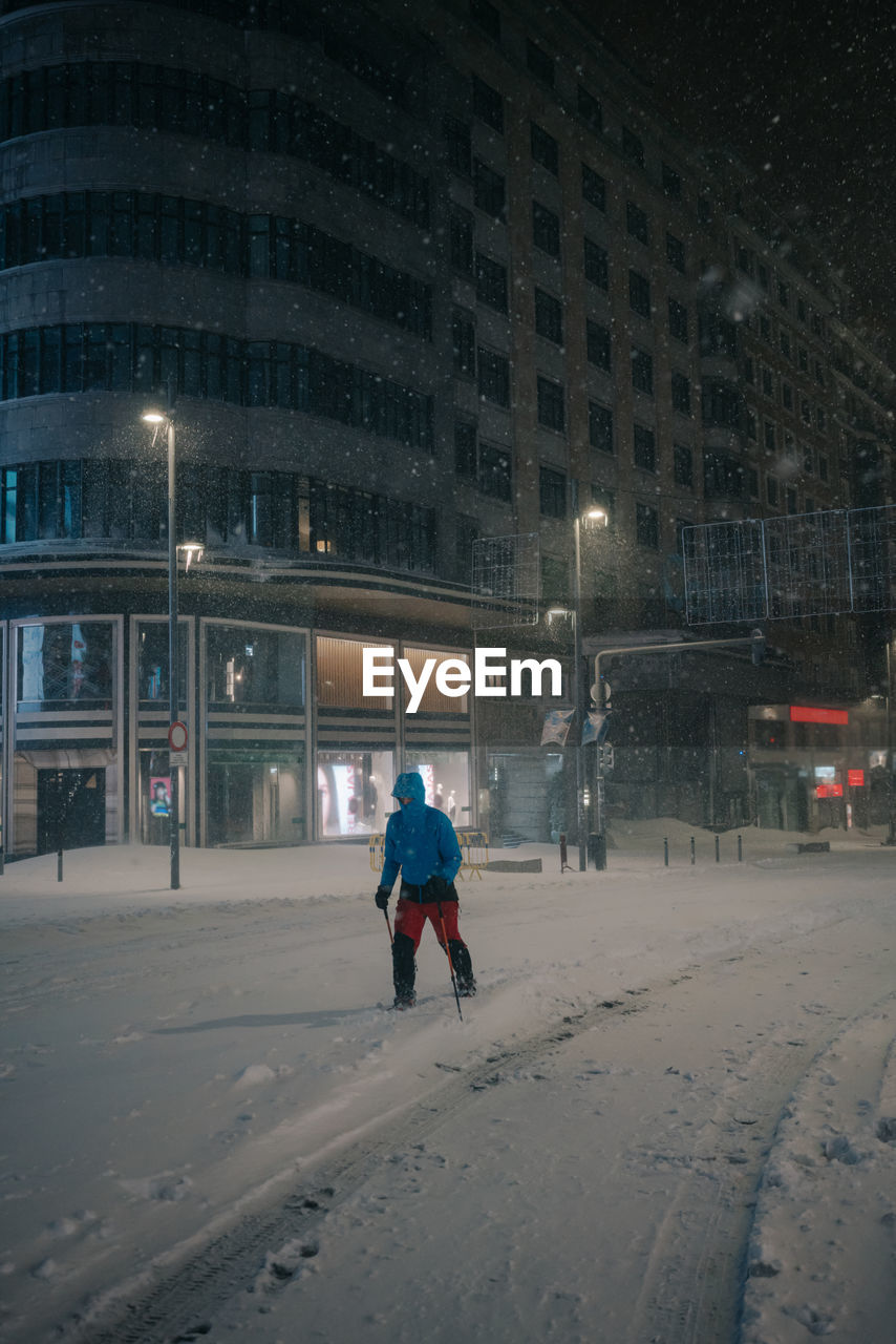 Anonymous male athlete in sportswear skiing on snowy road against building in evening during snowfall in madrid spain