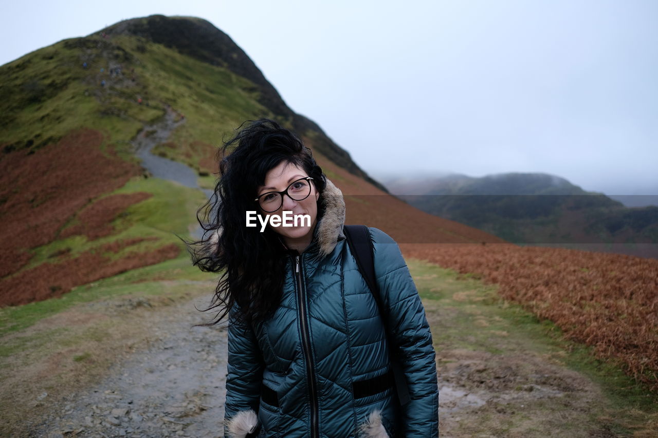 PORTRAIT OF SMILING WOMAN STANDING ON MOUNTAIN AGAINST SKY