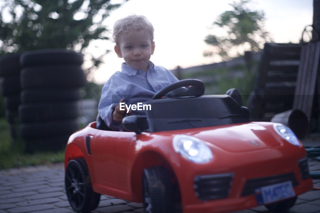 car, childhood, mode of transportation, vehicle, transportation, child, motor vehicle, one person, men, land vehicle, portrait, automobile, city car, day, sitting, casual clothing, driving, front view, sports car, emotion, happiness, smiling, leisure activity, outdoors, nature, standing, toy, looking, tree, innocence, looking at camera, lifestyles, clothing, wheel
