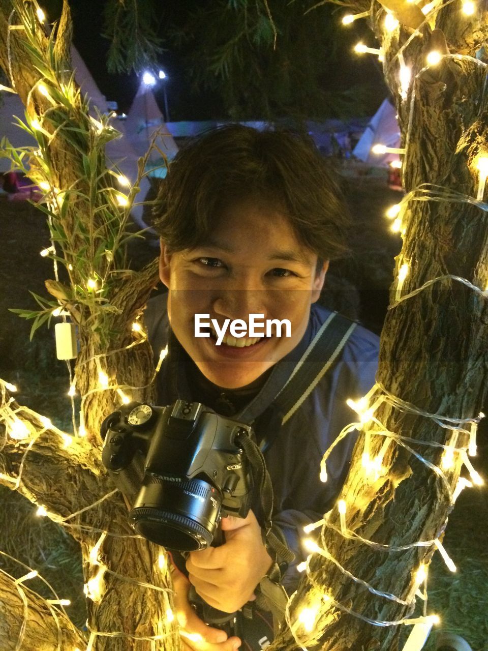 Portrait of smiling young man holding camera by illuminated tree